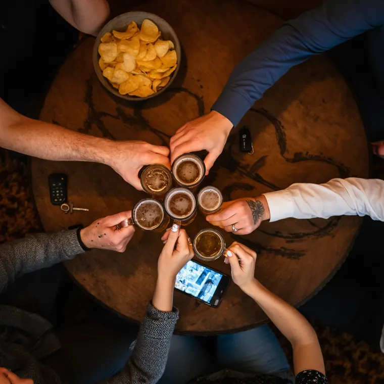 Six hands of different ages pushing schooners and middies together over a round pub table, shot from directly above.